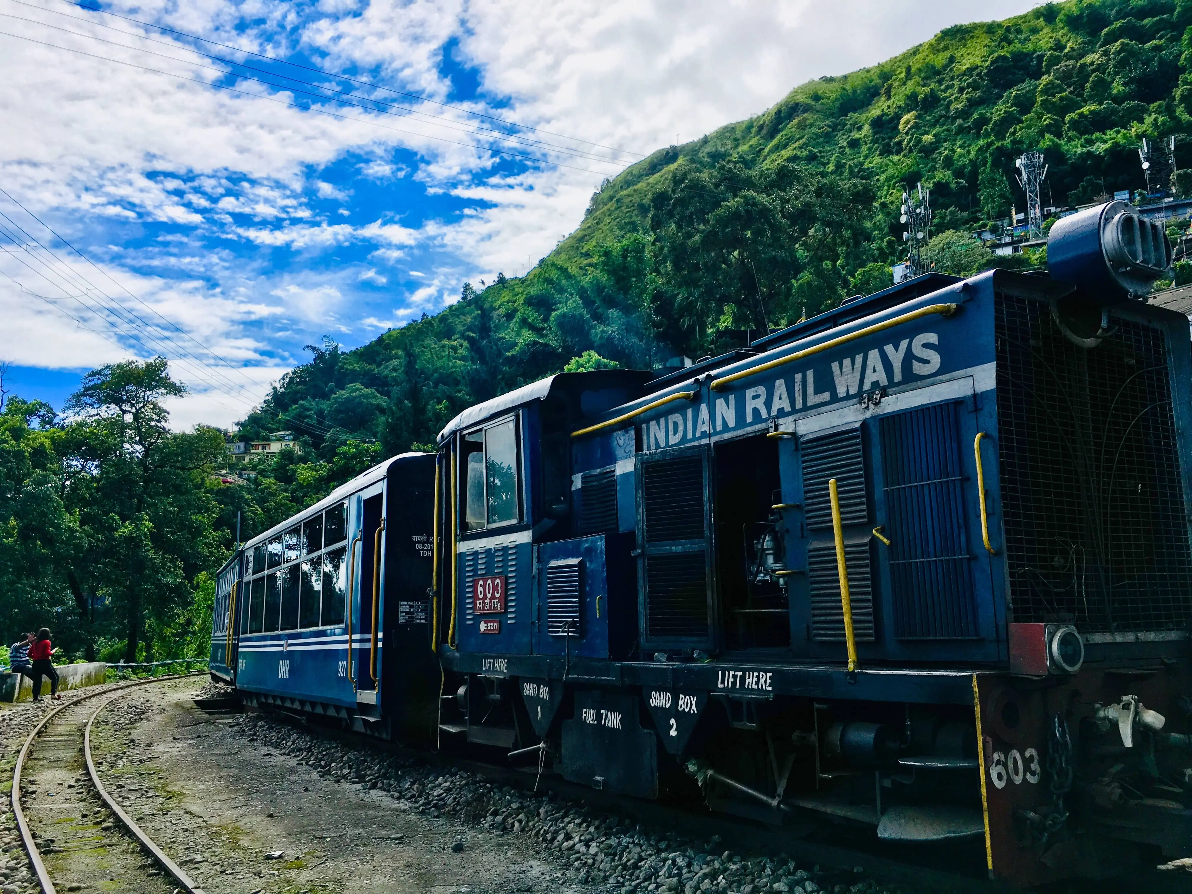 A blue train travelling along the Darjeeling Himalayan Railway through green hills and under a cloudy blue sky