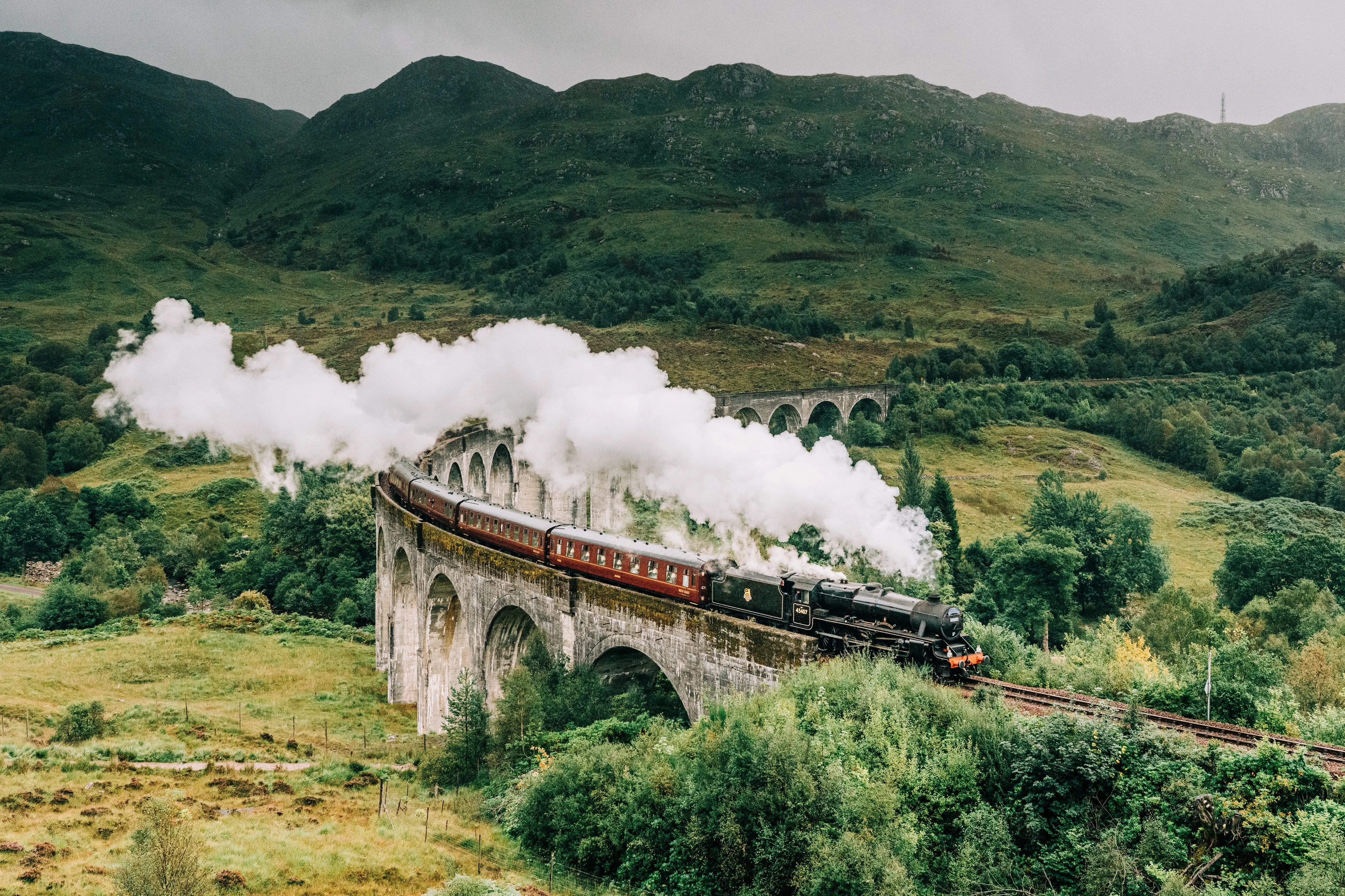 Train Network: A steam train travels along an aqueduct during the day as white steam plumes from its chimney.