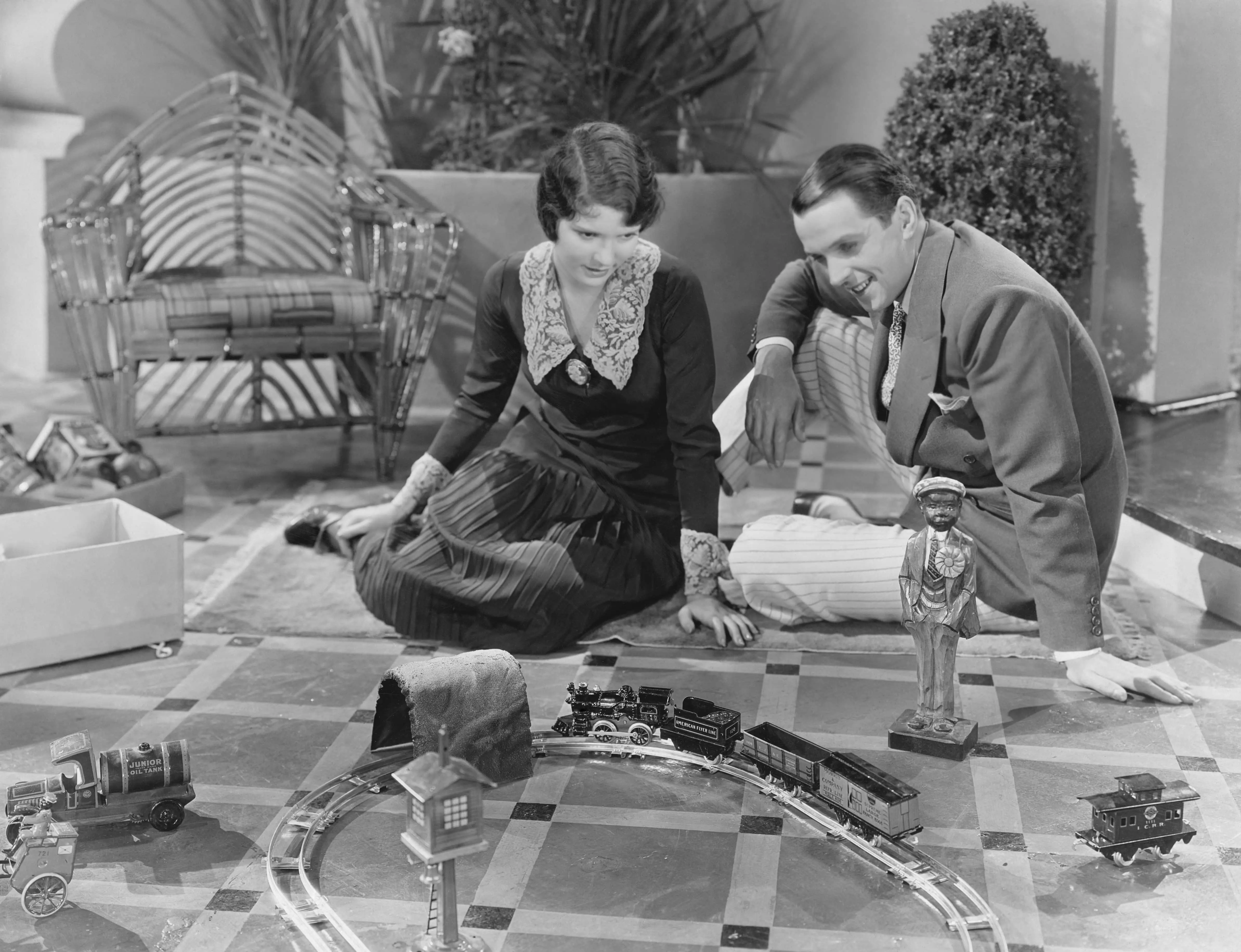 Model Trains: An old fashioned image of two young adults enjoying a model railway in their living room.